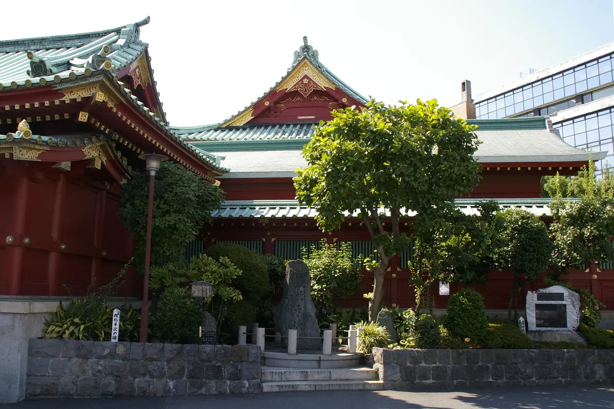 A vermilion shrine building with green roofs behind trees and a stone monument at the foot of a low wall at Kanda Myojin