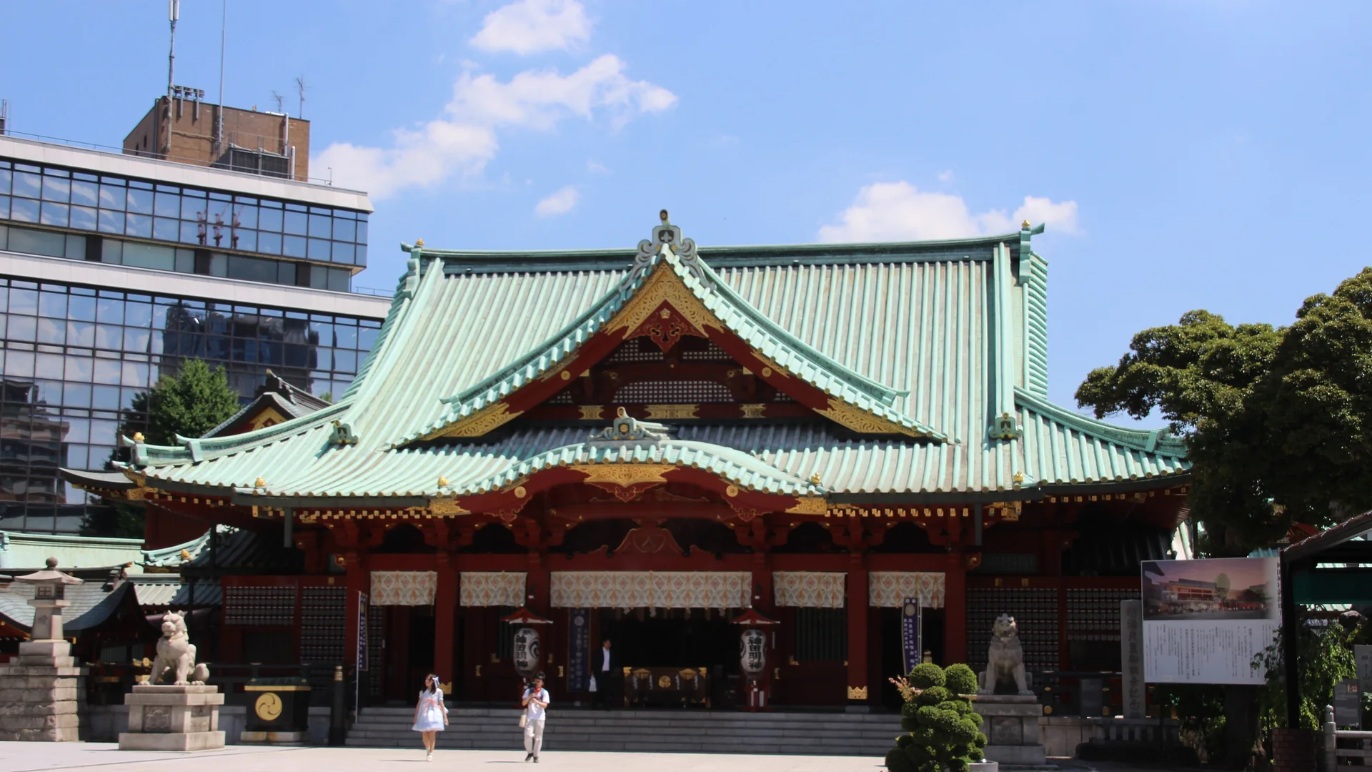 The vermilion main hall of Kanda Myojin shrine with a green tiled roof, seen across an open stone courtyard on a clear day
